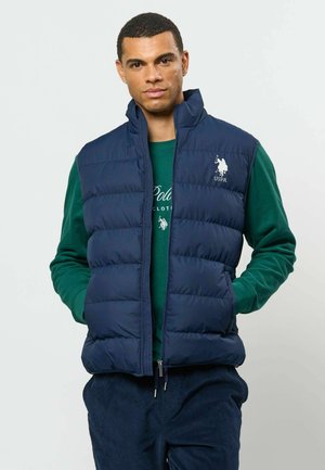 Homme portant une doudoune bleu marine sur une chemise verte à manches longues avec un logo blanc USPA, debout les mains dans les poches devant un fond uni.