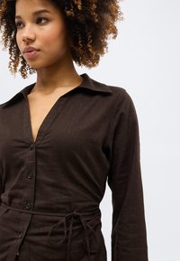 Νεαρή γυναίκα με σγουρά μαλλιά που φοράει σκούρο καφέ φόρεμα με κουμπιά, γιακά και ζώνη στη μέση, πάνω σε ανοιχτό φόντο.