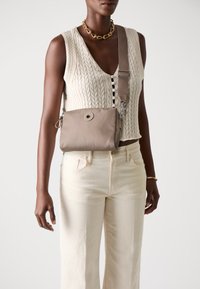 Borsa a tracolla beige in tessuto liscio, con dettaglio di bottone rotondo, tracolla regolabile e accessori in metallo argentato. Indossata sopra un top strutturato.