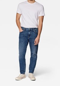 Mies, joka on pukeutunut yksinkertaiseen valkoiseen lyhythiharaiseen t-paitaan, sinisiin farkkuihin, käsi taskussa ja vaaleanharmaisiin kangaskenkiin, seisoo valkoista taustaa vasten.