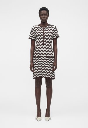 Čierna žena stojí, oblečená v hnedo-bielych šatách so vzorom cik-cak a krátkym rukávom, s prednými vreckami a bielymi topánkami so špicatou špičkou.