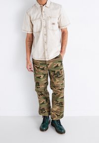 Beige overhemd met korte mouwen en knoopsluiting, voorzien van twee voorzakken, gecombineerd met groen en bruin camouflage cargo-broeken. Groene veterschoenen.