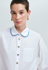 Camisa blanca con cuello redondeado que presenta un ribete azul, botones metálicos y un bolsillo en el pecho. La tela parece suave y ligera.