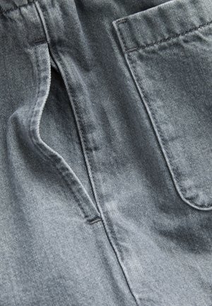 Țesătură din denim gri cu finisaj texturat, având un buzunar lateral și detalii de cusături. Materialul este ușor decolorat, evidențiind stilul său casual.