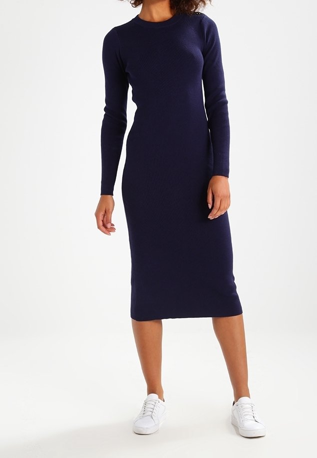 Femme portant une robe midi ajustée à manches longues bleu marine associée à des baskets blanches, debout devant un fond blanc uni.