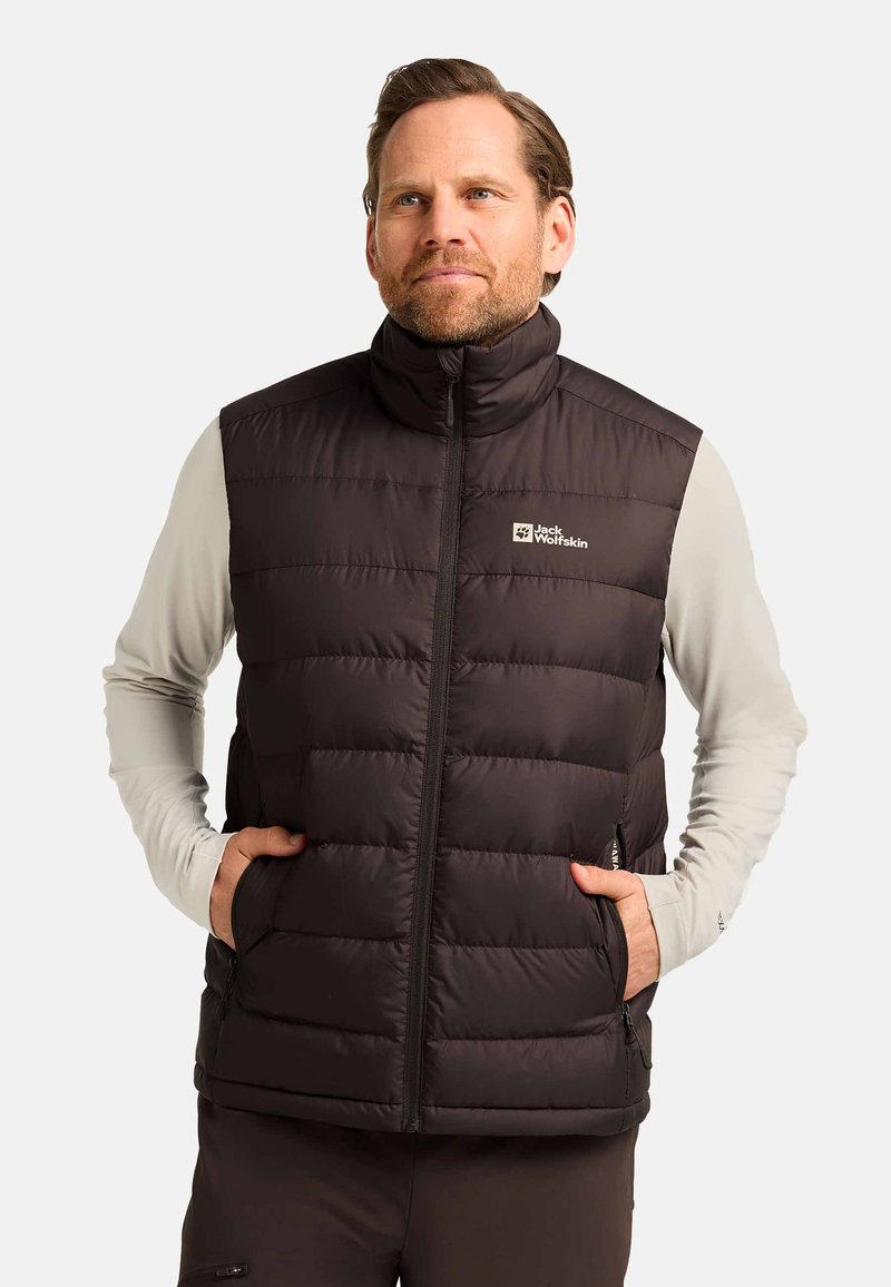 Man in een zwart gewatteerd mouwloos vest met rits en logo op de borst, met handen in de zakken, over een lichtgekleurde lange mouwen shirt, tegen een effen achtergrond.