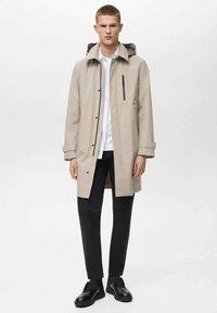 Cappotto impermeabile beige chiaro con cappuccio, maniche lunghe, tasca frontale con zip e chiusura a bottone, abbinato a pantaloni neri e scarpe.