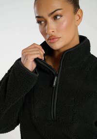 Pullover in pile nero con colletto alto e chiusura a half-zip, caratterizzato da una superficie testurizzata e da profili neri a contrasto lungo le cerniere.