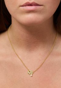 Goldene Kettenhalskette mit einem strukturierten Anhänger, der mit einer kleinen weißen Perle akzentuiert ist und elegant auf nackter Haut ruht.