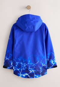 Veste à capuche bleue avec un motif d’éclairs électriques lumineux le long des manches et du bas, accrochée à un crochet en bois contre un mur blanc.