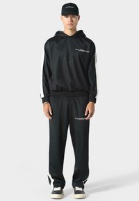 Tuta sportiva nera con accenti bianchi, cintura elastica, giacca con cappuccio, stampa del logo, indossata con sneakers nere e accessori.