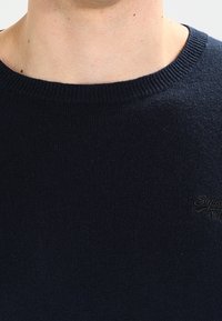 Tmavě modrý pletený svetr s kulatým výstřihem a jemným logem na hrudi. Měkká textura s žebrovanými manžetami.