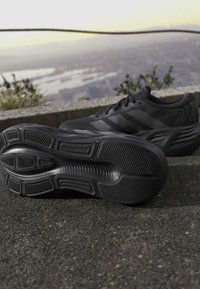 Pair of black athletic running shoes on asphalt near a stone ledge with a blurred urban landscape in the background.