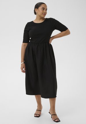 Femme portant une robe midi noire avec des manches au niveau du coude et une taille froncée, debout avec une main sur la hanche, portant des sandales noires à talons.