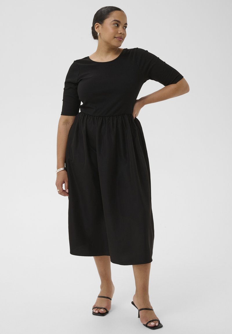 Femme portant une robe midi noire avec des manches au niveau du coude et une taille froncée, debout avec une main sur la hanche, portant des sandales noires à talons.