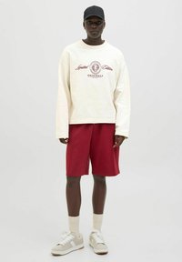 Sudadera color crema con gráfico rojo, pantalones cortos rojos y zapatillas gris. El conjunto presenta un corte holgado y pantalones cortos hasta la rodilla.