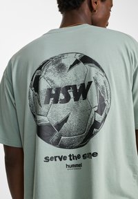 Ljusgrön bomullströja med en stor svart fotbollsgrafik och texten "HSW" samt "serve the game" nedanför. Korta ärmar, avslappnad passform.