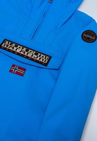 Veste imperméable bleue avec une poche avant, dotée d'un écusson logo noir et d'un petit drapeau norvégien. Texture lisse et design léger.