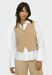 Beige fitted vest med fem knapper, båret over en hvid skjorte med krave. Den har en dyb V-udskæring og en cropped skæring. Egnet til lag-på-lag.