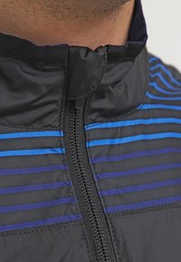 Chaqueta negra con rayas horizontales azules, con un alto cuello y una cremallera central para abrochar. Tejido suave y ligero.
