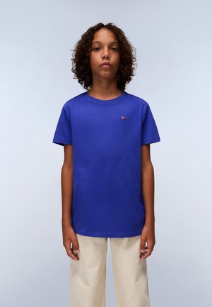 T-shirt en coton bleu royal avec un col rond et des manches courtes. Dispose d'un petit emblème de drapeau sur la poitrine. Associé à un pantalon large crème.