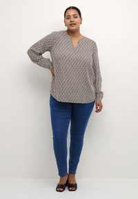 Langærmet bluse i sort og hvidt geometrisk mønster, V-hals, letvægtsstof. Båret med blå skinny jeans og sorte åbne sko.