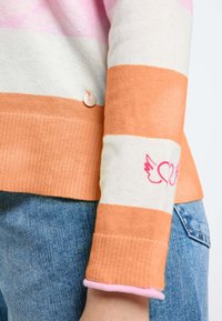 Gestreepte trui in roze, wit en oranje breiwerk. Heeft ribgebreide mouwen en een kleine borduursel van een hart en vleugel. Inclusief een metalen accent.