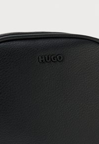 Čierna kožená taška s textúrovaným povrchom a vyvýšeným logom "HUGO" v čiernej farbe. Dizajn je kompaktný s zaoblenými hranami.