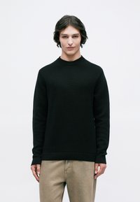 Νεαρός με μεσαίου μήκους σκούρα μαλλιά, φορώντας μαύρη μπλούζα με μακριά μανίκια και μπεζ παντελόνι, μπροστά σε έναν λευκό απλό φόντο.