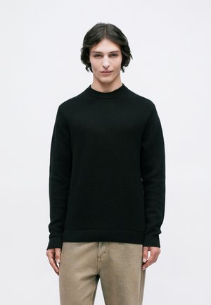 Jongere met halflang donker haar, gekleed in een zwarte lange mouwen trui en beige broek, tegen een eenvoudige witte achtergrond.
