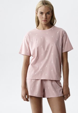 Blondynka w różowej koszulce z krótkim rękawem i dopasowanych szortach z małym, białym powtarzającym się wzorem tekstowym, stojąca na jednolitym jasnym tle.