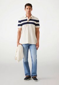 Beige poloshirt voor heren met marineblauwe horizontale strepen, gecombineerd met lichtblauwe spijkerbroek. Houdt een crème trui vast. Schoeisel: donkerbruine veterschoenen.
