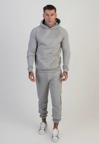 Sudadera con capucha gris claro y pantalones jogger a juego hechos de tela suave; cuentan con un bolsillo con cremallera, puños acanalados y detalles con el logo en ambas piezas.