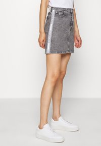 Gonna in denim grigio con vestibilità dritta, caratterizzata da un nastro con logo bianco lungo il lato, abbinata a sneakers bianche. Design pulito e casual.