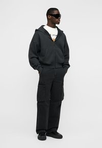 Homme portant un pantalon cargo noir surdimensionné, un sweat à capuche noir zippé, un t-shirt blanc à motifs, des baskets noires et de grandes lunettes de soleil noires, debout les mains dans les poches.