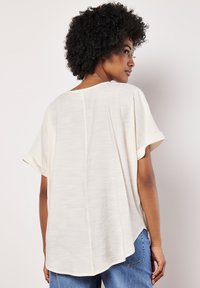 Blusa color crema a maniche corte con vestibilità rilassata, caratterizzata da una cucitura centrale sul retro e un tessuto morbido e testurizzato. Abbinata a jeans blu.