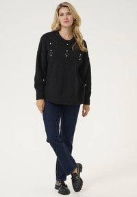 Melnā adīta džemperis ar teksturētām rakstām un pērļu akcentiem, savienots ar tumši zilām džinsiem un melniem loafer apaviem. Brīvas formas un ar apaļu kakla izgriezumu.