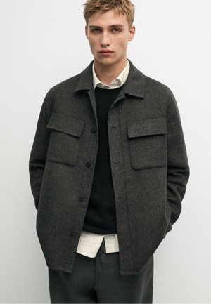 Homme blond porte une veste en laine gris foncé boutonnée par-dessus un pull noir et une chemise blanche à carreaux, debout les mains dans les poches.