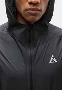 Κοντινό πλάνο ενός ατόμου που φοράει μαύρο μπουφάν με κουκούλα, με φερμουάρ μπροστά και λευκό τρίγωνο λογότυπο ACG στο στήθος.