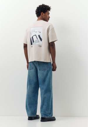 Uomo con maglietta beige chiaro con stampa grafica sulla schiena, jeans blu larghi e scarpe nere, in piedi di lato contro un muro bianco.