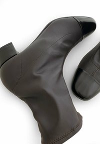 Botines de cuero marrón con una textura suave, punta redondeada, acentos de charol negro y un tacón robusto. Presentan costuras visibles.