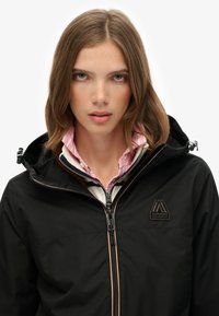 Veste noire imperméable avec fermeture éclair à l'avant et capuche ajustable. Présente un tissu texturé et un design de logo sur la poitrine.