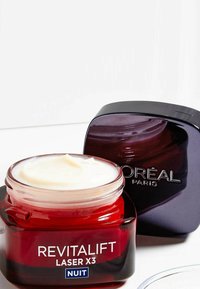 Bocal en verre rouge avec un couvercle transparent, étiqueté "Revitalift Laser X3 Nuit." Couvercle opaque violet foncé, texture crémeuse visible à l'intérieur.