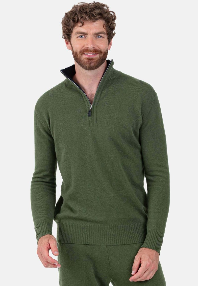Homme aux cheveux bouclés et barbe portant un pull à demi-fermeture éclair vert olive et un pantalon assorti, souriant devant un fond blanc.