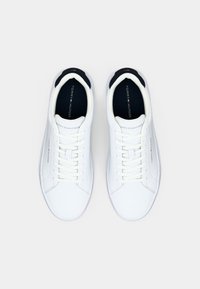 Paire de baskets basses blanches avec lacets blancs, doublure intérieure noire, et logo Tommy Hilfiger sur les semelles intérieures et les panneaux latéraux.