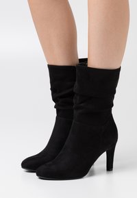 Botines de ante negros con un diseño arrugado, punta afilada y tacón apilado. Presentan una textura suave y sin herrajes o patrones visibles.