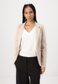Blazer beige con rever a punta, abbinato a un top bianco a scollo a V e pantaloni neri su misura. Texture liscia, design minimalista.