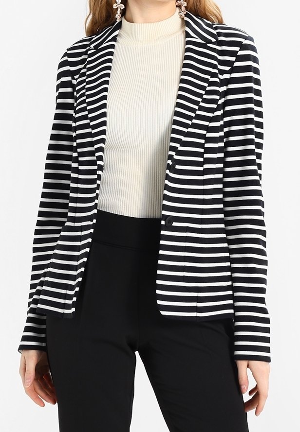 Femme portant un blazer à rayures horizontales noires et blanches sur un col roulé côtelé crème et un pantalon noir, debout avec les mains détendues.