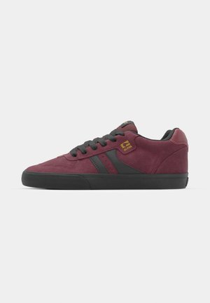 Burgundy suede sneaker with black accents, featuring a textured sole, round toe, and flat laces. Gold logo on the side.