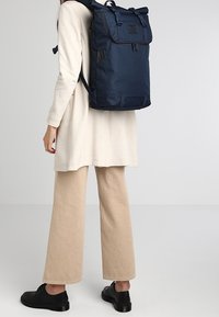 Námořnický modrý batoh s horní klopou, nastavitelné popruhy a obdélníkovým tvarem. Vyroben z odolné tkaniny, obsahuje přední zipovou kapsu a nášivku s logem.